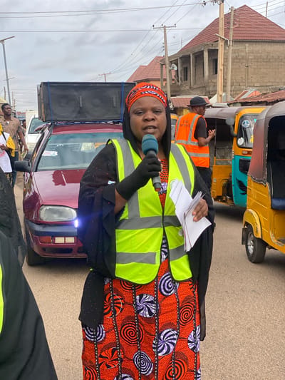 A medium shot captures a Black woman speaking into a microphone, positioned in front of a maroon car with large speakers mounted on its roof. She is wearing a bright orange and purple patterned headscarf and a reflective neon green vest over a black garment. She holds a stack of white papers in her left hand. Behind her, to her right, a man in an orange reflective vest labeled "CIVIL DEFENCE" stands near a yellow and teal auto-rickshaw. Further back, other auto-rickshaws and a building with a red tile roof are visible. The scene appears to be a street in Rigachikun, Nigeria, under an overcast sky.