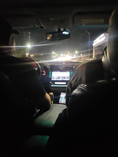 The image, taken at night, shows the interior of a vehicle, likely in Oskemen, Kazakhstan. A person, wearing a dark shirt and a black cap, is seated in the driver's seat, partially visible from the back. Their hands are on the steering wheel. The car's dashboard is illuminated, featuring a touchscreen display with a navigation interface. To the right of the driver, another person is seated, their head and shoulder visible from the back. The interior of the car is dark, with the primary light sources being the dashboard display and external streetlights visible through the windshield. Outside the vehicle, streetlights and building lights suggest an urban environment. The overall scene depicts a moment of travel at night, with the focus on the car's interior and the illuminated dashboard.