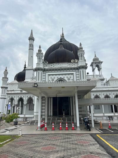 The image shows the exterior of Zahir Mosque in Alor Setar, Malaysia, on a cloudy day. The mosque is a grand structure with multiple domes and minarets, predominantly white with dark domes. In the foreground, a paved walkway leads to the entrance, bordered by red and white traffic cones. A motorcycle is parked on the right side of the walkway. A small tree in a pot is visible on the left side of the walkway. The architecture features intricate detailing, including arches and decorative patterns. The overall scene depicts a religious site with some temporary barriers and a parked vehicle.