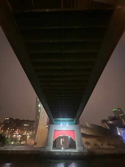 The image, taken at night in Bilbao, Spain, depicts the underside of a large, modern bridge. The bridge's structure, composed of dark, horizontal beams, dominates the upper portion of the frame. Beneath the bridge, the scene opens up to reveal parts of the cityscape. On the left, a cluster of illuminated buildings forms a dense urban background. In the center, a large, beige concrete pillar supports the bridge, featuring a prominent red, arched recess. To the right, the distinctive, metallic, curvilinear architecture of the Guggenheim Museum Bilbao is visible, with its reflective surfaces catching the ambient light. The foreground shows a dark body of water, likely the Nervión River, with subtle reflections of light on its surface. The sky is overcast, creating a hazy, diffused lighting condition. There are no discernible people or active interactions depicted in the scene.