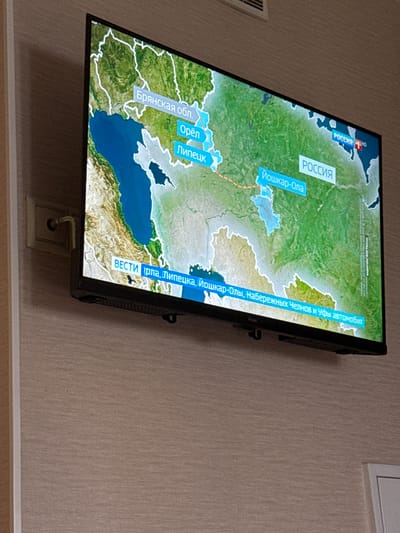 A widescreen television displays a map of Russia with several cities labeled, including Bryansk Oblast, Oryol, Lipetsk, and Yoshkar-Ola. Text overlays indicate the channel "Rossiya" with an "HD" logo. Another text overlay at the bottom of the screen reads "VESTI" followed by a partial sentence in Russian, which translates to "reports from Iran, Lipetsk, Yoshkar-Ola, Naberezhnye Chelny and Ufa by car." A power outlet and cable are visible to the left of the television, mounted on a beige textured wall. The TV is positioned at a slight upward angle. The image is taken from a slightly elevated perspective.