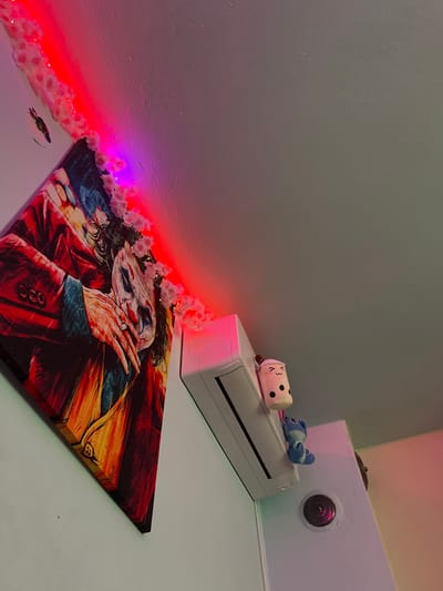 The image depicts an indoor room with elements of decoration and utility. A large canvas print of a character resembling the Joker, featuring a painted face, red suit, and visible hands, is prominently displayed on a light-colored wall, oriented diagonally. Along the intersection of the wall and ceiling, a string of LED lights transitions from red to purple, entwined with numerous small, white artificial flowers, which also extend onto the upper right edge of the artwork. A small, dark, reflective silhouette, possibly a bird, is affixed to the wall above the artwork. To the right of the artwork, a white split-unit air conditioner is mounted high on the wall. Resting on or attached to the air conditioner are two plush toys: one depicting a white boba tea cup with a smiling face, and another smaller, blue, cartoonish figure resembling Stitch, hanging upside down. Further to the right, a circular, dark object, resembling a camera lens or peephole, is embedded in a vertical, light-colored surface, reflecting a reddish tint. The ceiling above the air conditioner is light gray, while the wall section to the far right appears light blue, blending into pinkish-purple illumination. No individuals are present. The specific location in the city of Debelt, Bulgaria, cannot be determined from the visual information provided.
