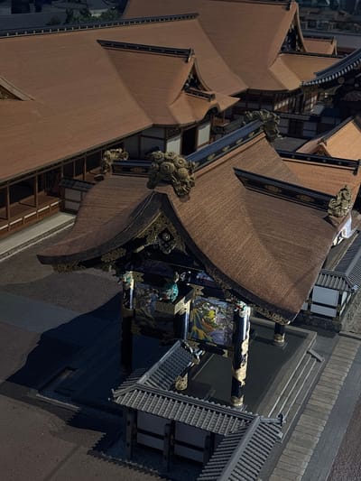 This is an aerial, high-angle, eye-level, exterior, full shot of the National Theatre in Chiyoda City, Tokyo, Japan. The theatre, built in 1966, features a traditional Japanese architectural style with multiple tiered, dark brown roofs. The main building is accompanied by smaller structures, also with similar roofing materials. Decorative golden ornamentation is visible on the roof edges and on pillars supporting the main structure. The building complex is surrounded by grey pathways and some landscaping elements, including trees visible in the background. The lighting suggests it is either early morning or late evening, with some shadows cast across the complex. There are no people visible in the scene.