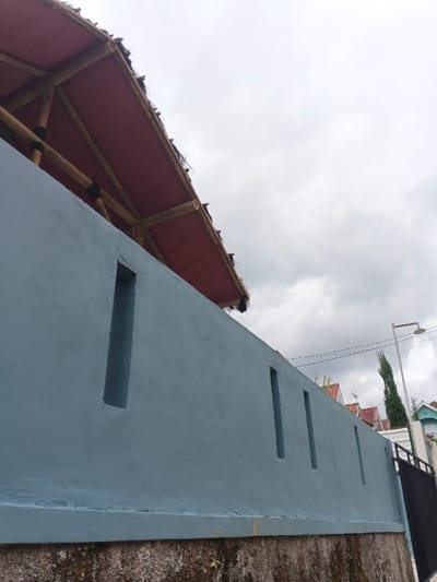A blue wall with vertical rectangular openings spans the foreground. Mossy rock forms the base. Above the wall, a red-roofed structure with bamboo supports is partially visible. The background shows glimpses of houses with red roofs, a tall, slender tree, and a streetlight against a cloudy sky. A black fence stands at the right edge of the image.