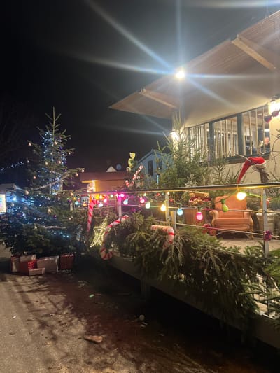 A building exterior is decorated for Christmas. Christmas trees with ornaments and string lights are prominent. A balcony above features a plush chair, a Santa figure, and more decorations. Candy canes and gifts surround the trees. A streetlight shines intensely from above. The overall scene is festive and illuminated against the night sky.