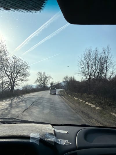 The image provides a driver's perspective from a vehicle on a road in Gorna Verenitsa, Bulgaria, under a clear, sunny sky. The dashboard is visible in the foreground, partially covered by a light-colored mat with two pieces of white tape adhered to it. The windshield frames a two-lane asphalt road stretching forward and curving slightly to the right. Sunlight creates bright reflections and prominent shadows from trees on the road surface. A light-colored minivan or SUV travels ahead in the same direction. To the left, several large deciduous trees, bare of leaves, line the road, with distant hills or mountains visible on the horizon. To the right, a verge with dry vegetation and concrete road barriers runs parallel to the asphalt, backed by more leafless trees and bushes. The blue sky above displays three white contrails; two are parallel, and a third intersects them. A small, dark, indistinct airborne object is present in the mid-right portion of the sky.