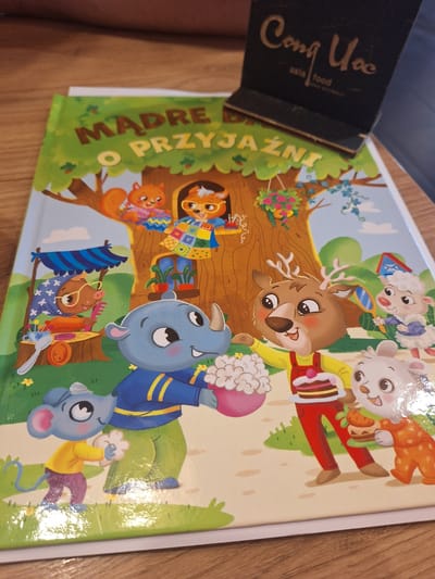 This is a close-up, top-down shot of the cover of a children's book. The book's title, "MADRE O PRZYJAŹNI," is prominently displayed in large, green, three-dimensional letters. The cover features a colorful illustration with various anthropomorphic animal characters and a tree as the central element.
The subjects include a deer, a fox, a pig, a sheep, a goat, and mice, all depicted in a whimsical, cartoon style. They are engaged in various activities, suggesting a narrative of friendship and perhaps celebration, with one character holding a cake and another a flower bouquet. The setting appears to be a whimsical forest or garden. The overall emotion conveyed is cheerful and playful.
Notable details include the bright, vibrant colors of the illustration, indicating it's likely daytime. A black object with the text "Cong Uoc asia food" is partially visible in the top left corner, leaning against the book. The image appears to be taken in a home or casual setting, with a wooden table surface visible beneath the book. The context provided places this scene in Mińsk Mazowiecki, Poland.