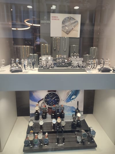 The image shows a display window of a jewelry store, likely located in Rosenheim, Germany, given the presence of a "MADE IN GERMANY" label and the style of the watches. The display is divided into two main sections. The upper section showcases men's jewelry, including necklaces, bracelets, and rings, displayed on grey stands. A sign above reads "HERREN-SCHMUCK" (Men's Jewelry) and "OROVIVO 1856." The lower section features a collection of wristwatches, with several models arranged on tiered black stands. A large graphic behind the watches depicts a Zeppelin airship. The watches are varied in style, with leather and metal straps, and some have visible price tags. The overall environment appears to be a well-lit retail setting. There are no people visible in the image.