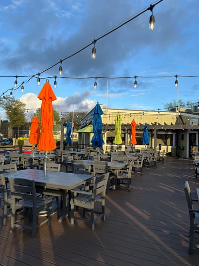 The image shows an outdoor patio area in Laconia, United States of America. The patio is constructed of dark brown wooden planks and is furnished with numerous gray picnic tables and chairs. Each table has a closed, colorful umbrella, with a repeating sequence of blue, orange, and green. A string of exposed bulb lights is strung above the tables. The sky above the patio is blue with some dark, ominous clouds. In the background, trees and other buildings are visible. There are no people present in the image.