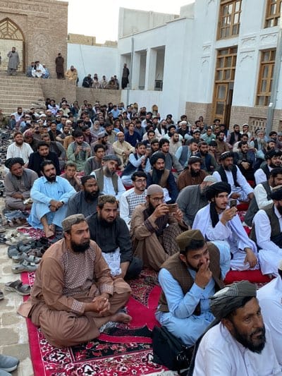 The image depicts a large gathering of men, predominantly dressed in traditional Afghan attire, seated on rugs in an outdoor area. They appear to be attending an event or religious service. The setting seems to be in the city of Kandahar, Afghanistan, suggested by the architectural style of the buildings in the background, which feature earthy tones and intricate stonework, characteristic of Afghan and Islamic architecture. Some men are looking towards a central point not visible in the frame, while others are engaged in conversation or looking at their phones. Shoes are visible, neatly placed on the ground in some areas, indicating a respectful atmosphere. The overall scene conveys a sense of community and religious observance.