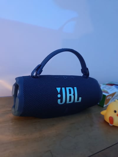 A blue JBL speaker with a handle is lying on a wooden surface. Next to the speaker, there is a small yellow chick toy and a partially visible box. The background is a light blue wall. The scene is lit with a cool light. No location or city is evident in the image.