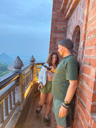 A man and a woman are standing on a brick balcony overlooking a landscape at dusk. The man is wearing a green t-shirt, green shorts, and a black baseball cap. He has a beard and is looking down at his phone, which he is holding in his right hand. A watch is visible on his left wrist. The woman is wearing a light-colored crop top and green shorts. She has sunglasses perched on her head and is also looking at a phone, which she is holding in her left hand. Her right arm is extended slightly as if gesturing or reaching out. Both individuals appear relaxed and engaged with their devices.
The setting is characterized by a brick wall with an arched opening, suggesting an old structure or architectural feature. Below the balcony, a staircase with ornate stone carvings and metal railings is visible, illuminated by warm artificial lights. The landscape in the background features rolling hills covered in lush greenery, with a hazy sky indicating either evening twilight or a slightly overcast condition. The overall atmosphere seems peaceful, with the soft light suggesting it is either early morning or late evening. The location context provided places this scene in Tây Hoa Lư Ward, Vietnam, hinting at a possibly historical or scenic tourist spot. No visible text is present in the image.