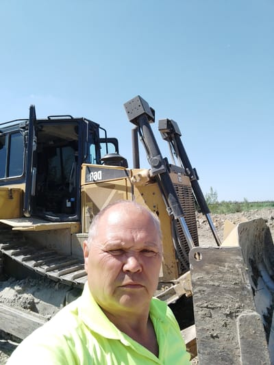 A middle-aged Caucasian man with gray hair and a serious expression is taking a selfie in front of a yellow bulldozer. He is wearing a bright yellow-green polo shirt. The bulldozer has its doors open, revealing the interior of the cab. The tracks of the bulldozer are visible in the foreground, and a large blade is positioned to the right of the man. The background shows a construction site with dirt piles and some sparse vegetation under a clear blue sky. A crescent moon is faintly visible in the sky. The image was taken outdoors during the day, likely in a rural or industrial area. The location is identified as Rozdil, Ukraine.