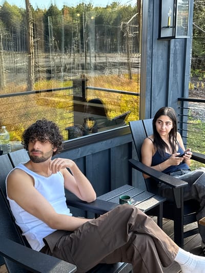 An outdoor scene in Otego, United States, depicts two individuals seated on a wooden deck. On the left, a male with dark curly hair and a beard, wearing a white tank top and dark brown pants, sits in a dark grey Adirondack-style chair, looking towards his left. A clear plastic water bottle rests on the armrest of his chair. To his right, a female with long dark hair, wearing a dark blue spaghetti strap top and light-colored jeans, occupies a similar dark grey Adirondack-style chair. She holds a smartphone in both hands and looks towards the left. A small integrated table between the chairs holds a dark green mug. The deck features dark grey siding and horizontal metal railings. Behind the subjects, a large window reflects a natural wetland environment characterized by numerous thin, bare tree trunks standing in water or wet ground, surrounded by yellow-brown foliage, with a denser green forest visible in the background under a clear sky. A black metal outdoor wall sconce is mounted on the exterior siding to the right of the window.