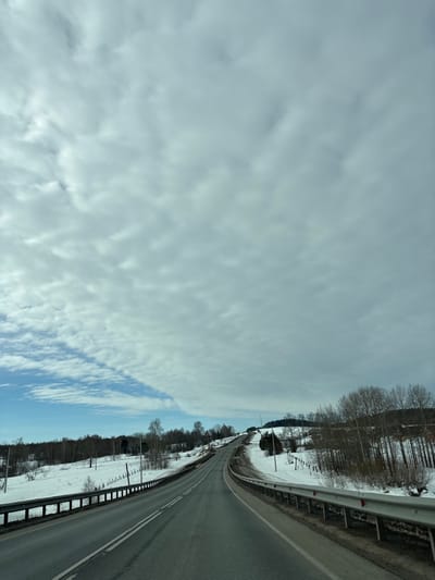 This image depicts a two-lane asphalt road ascending and curving slightly to the right through a snow-covered, rural landscape in Vakhrino, Russia. The sky is largely overcast with dense altocumulus clouds, though a section of blue sky with scattered white clouds is visible in the upper left. Metal guardrails line both sides of the road, which features a solid double white line separating the lanes. Snow blankets the ground and hillsides. Bare deciduous trees and some scattered conifers populate the slopes. Utility poles with lines are visible along the left side of the road, and a wire fence runs parallel to the road on the left. No individuals or vehicles are present in the scene.
