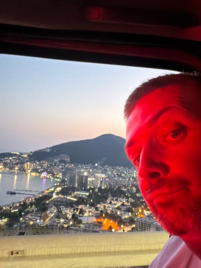 A close-up, selfie-style photograph captures a man's face, bathed in red light, from the chest up. He is looking towards the camera with his right eye visible and his mouth slightly open. The background reveals a panoramic view of a coastal city at dusk. The city lights twinkle, indicating a densely populated area with buildings and structures cascading down a hillside towards a body of water dotted with boats. A large, dark hill or mountain dominates the far background, silhouetted against a pale sky. A sliver of a crescent moon is faintly visible in the upper left quadrant of the sky. The perspective suggests the photo was taken from a high vantage point, possibly a vehicle, given the red interior visible at the top of the frame and the railing in the foreground. The specific location is identified as Košljun, Montenegro.