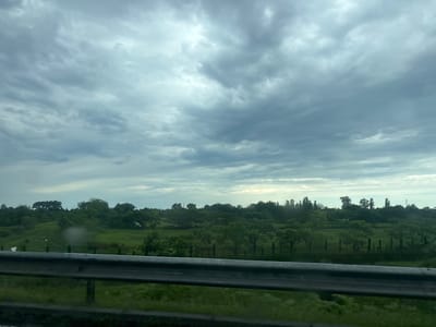 The image shows a landscape scene with a cloudy sky. In the foreground, there is a metal guardrail running horizontally across the lower part of the frame. Beyond the guardrail, there is a green field with some scattered trees and foliage. The sky is overcast with thick, gray clouds. The scene appears to be an outdoor environment, possibly a rural area or a roadside view. The location is specified as Nakaidzeebi, Georgia. There are no people or animals visible in the image, and no specific actions or interactions are depicted. The overall impression is one of a natural landscape under a somber sky.