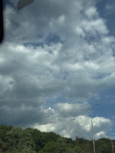 A roadside scene captures a cloudy sky over a dense, green forest. A tall, rusty lamppost stands prominently in the foreground on the right, its light fixture extending horizontally. In the distance, several cell towers are visible above the treeline. The weather appears overcast, with dramatic cloud formations dominating the sky. The image is taken from a low vantage point, likely from a vehicle, as suggested by a dark, blurred object in the upper left corner, possibly part of the car's interior.