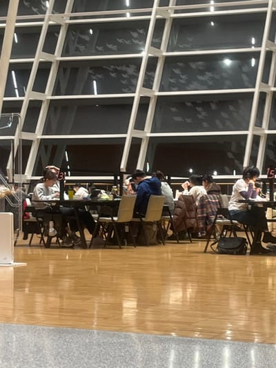 The image displays an indoor scene within a large, modern building in the city of Yanqiao, China. The background features a tall, multi-paneled glass wall structured by a prominent white grid framework. Numerous light-colored, stylized bird-like silhouettes are applied to the glass panes, creating a patterned effect. Beyond the glass, the exterior appears dark, with faint illumination in the distance. The foreground consists of a reflective light-brown wooden floor, which transitions to a gray textured floor surface at the bottom edge of the frame.
In the midground, a row of dark rectangular tables is set up with light-colored folding chairs. At least eight individuals, appearing to be young adults, are seated at these tables. Most are dressed in casual long-sleeved attire, including sweaters and a blue hooded jacket. A brown puffer jacket and a plaid shirt are draped over two chairs. Individuals are focused downwards, engaged in solitary activities such as working on laptops or reviewing materials. Laptops, books, and water bottles are visible on the tables. One person on the far right wears over-ear headphones. A black messenger bag rests on the floor next to a chair on the right. A small white rectangular device stands on the floor in the left foreground. Numbered signs, specifically "3" and "15", are visible above the seated individuals, indicating specific sections or seating arrangements. No direct interactions among the individuals are discernible.