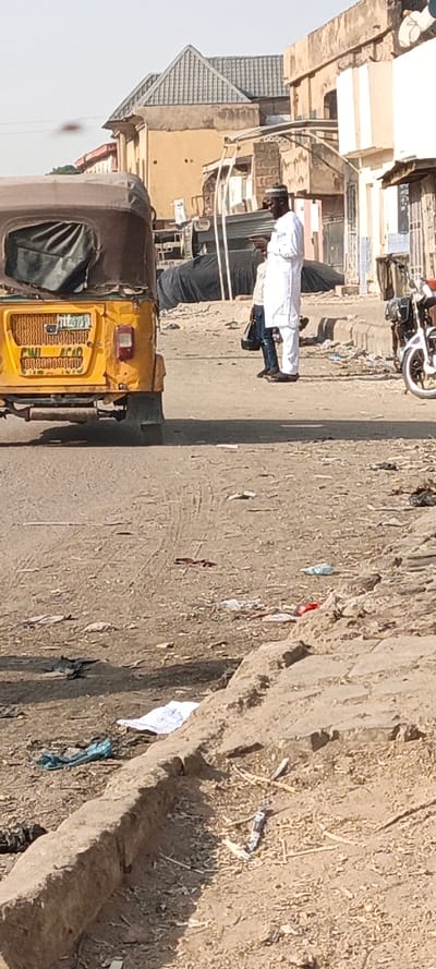 The scene is an outdoor, daytime view in Kaura Goje, Nigeria, showing a dusty, unpaved road and surrounding structures. On the left, the rear of a yellow tricycle with a brown fabric canopy is visible, displaying a license plate "CWR 4731" and additional Arabic script. The road surface is uneven, marked with tire tracks, and heavily littered with various debris including plastic bags, paper, and miscellaneous refuse.
In the mid-ground, two males are standing on the dirt shoulder of the road. The man on the right, dressed in a long white traditional robe and a white kufi cap, stands facing left with his right arm raised in a gesturing motion. Partially obscured behind him, a second male is visible, wearing blue jeans and holding a dark bag in his left hand. To the far right, the front wheel and partial frame of a white motorcycle are visible.
The background is populated with several buildings of varying construction. A prominent structure features light-colored walls and a distinct, grey-patterned gabled roof. Other buildings appear to be in various states of finish or disrepair, with some showing exposed brickwork and simplified architectural forms. A concrete curb or drainage edge runs diagonally across the lower right foreground.
