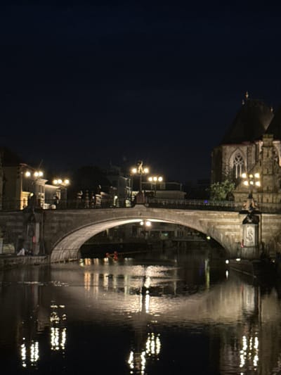 An illuminated stone bridge arches over a dark body of water in Ghent, Belgium, at night. The bridge is lined with streetlights, casting a warm glow on the stone structure and reflecting in the water below. Buildings, also illuminated, are visible on either side of the bridge, with a prominent church spire rising on the right. The water reflects the lights from the bridge and buildings, creating a shimmering effect. The sky is dark and clear, indicating a nighttime scene.