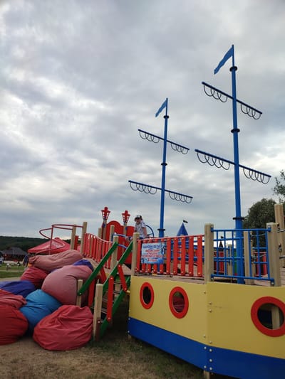 In Shcherbani, Ukraine, a vibrant playground designed to resemble a pirate ship is depicted under a cloudy sky. The main structure features large blue masts with horizontal spars and rigging, suggesting a nautical theme. Below the masts, a wooden deck area is surrounded by colorful railings in red and blue. Red beanbag chairs are piled near the entrance of the playground. A young person wearing a patterned t-shirt and a baseball cap is visible on the upper deck of the ship structure. Signs are posted on the railing, including one with Cyrillic text that appears to be a warning or instruction for using the attraction. The background shows greenery and some buildings, suggesting an outdoor recreational area. The ground is covered with dry grass and dirt.
