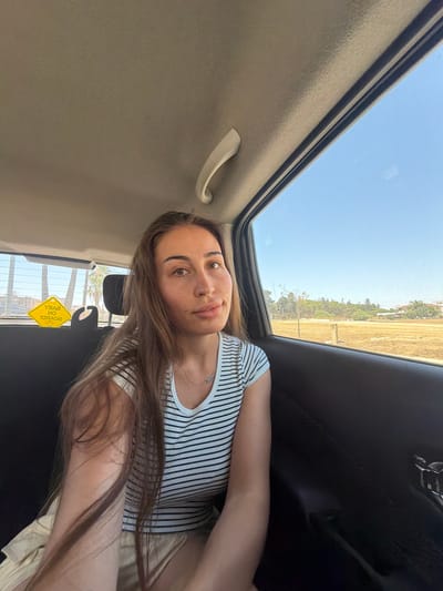 A young woman with long, brown hair is seated in the back of a car, looking towards the camera. She is wearing a black and white striped shirt and a light-colored skirt. A yellow "Baby on Board" sign is visible on the car window behind her. The car is traveling through a sunny, semi-arid landscape with scattered trees and buildings in the distance, consistent with the outskirts of Larnaca, Cyprus. The sky is clear and blue.