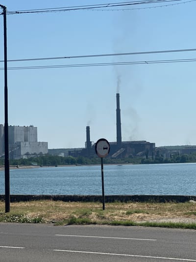 The image shows a wide view of an industrial complex situated beside a large body of water, likely a reservoir or river. The complex features several large structures, including a prominent, tall, smokestack emitting visible smoke, and a very large, pale, rectangular building with numerous windows and conveyor belts visible on its exterior. Another section of the industrial plant, appearing older and darker in color, is also visible, with a more complex arrangement of buildings and structures. The water in the foreground is calm and reflects the clear, blue sky. The shoreline is lined with green trees and vegetation. In the immediate foreground, a paved road runs horizontally across the bottom of the image, with a grassy verge and a dark pole with electrical wires running overhead. A circular road sign with a red border and a black car silhouette on a white background is posted on a metal pole in the middle ground. The overall scene suggests an active industrial area, likely located in a setting with natural water resources, consistent with the TPP Galabovo industrial site in Galabovo, Bulgaria.