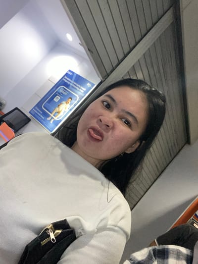 The media file is a selfie taken by a young woman with dark hair who is sticking her tongue out at the camera. She is wearing a white long-sleeved shirt and has a black bag with a gold zipper over her shoulder. The background appears to be indoors, possibly a waiting area or office, with a gray metal shutter in the upper part of the image. To the left, there is a blue poster with text and an image of a person, and a white light fixture is visible on the ceiling. To the right, another person wearing a blue and white plaid shirt is partially visible. The lighting suggests it could be daytime or an indoor setting with artificial lights. The woman's expression conveys a playful or humorous emotion. There is text visible on the blue poster, but it is not legible enough to be transcribed. The location context is Watampone, Indonesia.