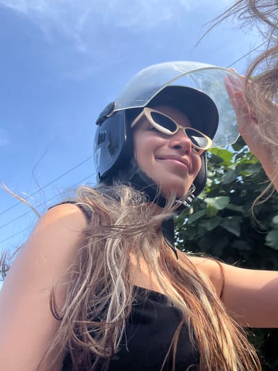 In Batawala, Sri Lanka, a young woman wearing a black helmet and cat-eye sunglasses smiles while holding up the visor of her helmet. Her long, sun-kissed hair, a mix of blonde and brown, blows in the breeze around her face. She is wearing a black top, and her right arm is visible, as is part of her left shoulder. Behind her, lush green foliage is visible, and the bright blue sky is clear with a few wispy clouds. A few power lines are also present in the background.