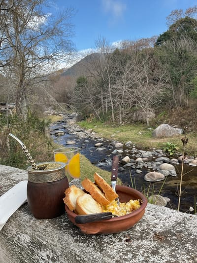An overhead, close-up, eye-level shot captures a rustic breakfast spread set against a scenic natural backdrop. In the foreground, a brown gourd filled with yerba mate and a metal bombilla sits to the left, next to a glass of orange juice adorned with orange slices. To the right, a clay bowl holds scrambled eggs and toasted bread slices, with a fork and knife resting in them. A white napkin is tucked beneath the gourd. The breakfast is positioned on a rough-hewn stone ledge overlooking a shallow, rocky stream that flows through a wooded area. Bare trees and green foliage dot the landscape, with a gentle slope of the terrain leading up to a hazy mountain in the distance. The sky above is a clear blue with a few wispy clouds. The scene suggests a tranquil morning meal in a natural setting, likely in the vicinity of El Rodeo, Argentina, known for its picturesque mountainous terrain and streams. There are no people or animals visible in the image.