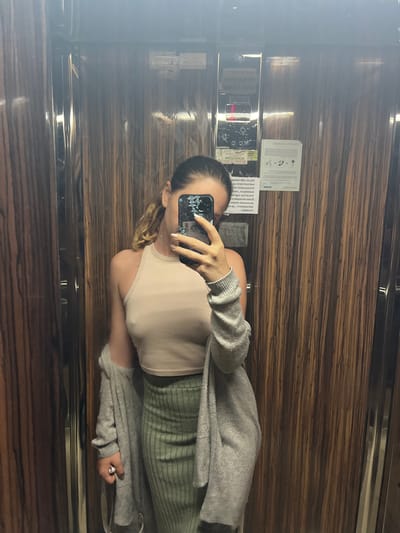 A young woman with blonde hair tied in a ponytail is taking a mirror selfie in an elevator. She is wearing a light beige halterneck top and a long, light grey cardigan draped over her shoulders. Her lower body is clad in a green, ribbed, knee-length skirt. The elevator walls are made of dark wood paneling with vertical grain. On the elevator wall to her right, there is a control panel with illuminated buttons and some signage. The overall lighting is dim, creating reflections on the elevator's mirrored surfaces. The image does not provide any specific visual clues to indicate the location is in Lozenets, Bulgaria.