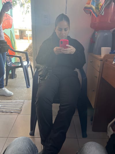 This image depicts an indoor setting in Quilmes Oeste, Argentina, with a doorway providing a view to an outdoor residential area during daylight. The central subject is a female, seated on a dark plastic chair, facing slightly left. She has dark hair, wears a black long-sleeved top and black trousers, and a black holster containing a firearm is visible on her right hip. She holds a bright pink smartphone in both hands, actively engaged with the device, looking down at its screen.
To the left, partially visible, stands another individual wearing blue jeans, white sneakers, and a light-colored fanny pack, holding a dark smartphone. The immediate indoor floor is tiled. Against the right wall, a light-colored wooden dresser or cabinet holds a red and grey water dispenser with an inverted water bottle, a white mug, and a folded patterned cloth. A white electrical outlet is mounted on the wall to the subject's left.
Through the doorway, an outdoor concrete surface is visible, along with another dark plastic chair. A wall adorned with orange and green mural elements is present on the left side of the outdoor view. Beyond this, residential buildings and some vegetation are visible in the background. The legs of a third individual, clad in light-colored jeans, occupy the immediate foreground, indicating the photographer's perspective.