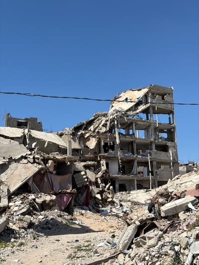 The image displays a scene of widespread destruction in Jabalia, Palestinian Territory, under a clear blue sky. The central focus is a heavily damaged multi-story concrete building, with its upper floors significantly collapsed and structurally compromised. Concrete slabs, rebar, and debris are extensively scattered around and within the remaining framework of the building. Multiple floor levels are visible, with internal rooms exposed to the exterior, indicating severe structural failure.
The ground in the foreground and surrounding the central building is covered in a dense field of rubble, composed of broken concrete, twisted metal, and assorted building fragments. A dirt path is visible through the rubble in the immediate foreground, leading towards the base of the damaged structure. Amidst the debris, particularly to the left of the central building, small makeshift structures or coverings made of draped fabric (appears reddish-brown and beige) are present. Overhead, a single black electrical or communication wire stretches horizontally across the upper third of the frame. No individuals or active movements are discernible within the scene. The overall environment suggests a site of extensive demolition or impact.