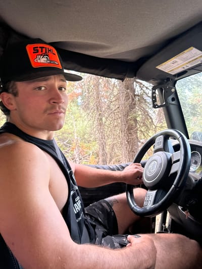 A single adult male is depicted in the driver's seat of a black Jeep vehicle within a wooded environment in Crystal Lake, United States. He possesses a light complexion, dark hair, and a visible mustache. He wears a black baseball cap featuring an orange "STIHL" logo with a chainsaw graphic, a black sleeveless shirt displaying partially legible white text ("CLAY...KING"), and camouflage-patterned shorts. A tattoo is visible on his right thigh. His right hand rests on the black and silver steering wheel, which bears the "Jeep" logo, while his left hand is placed on his left thigh. He is looking directly at the viewer with a neutral facial expression. The vehicle's interior features a fabric roof lining, a passenger-side sun visor with warning labels, and an aftermarket mirror assembly mounted on the passenger-side A-pillar. The background reveals a dense forest composed of numerous tall, slender trees, many appearing bare or with sparse foliage, indicating a natural, possibly off-road, setting under bright daylight.