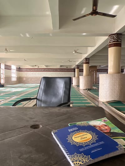 The image captures an interior view, possibly within a mosque. In the foreground, a gray plastic table holds two books. The top book has a blue cover with the names "Hayatudeen & A'isha," and the date "SUN. 8TH FEBRUARY, 2026," indicating a possible wedding or celebration. Underneath it is a book with a man wearing a white cap on the cover.

A black office chair is positioned beside the table. Beyond the table and chair, the architectural elements of the mosque are visible. There are green patterned walls, along with a series of columns or pillars with decorative tops. Ceiling fans are spaced along the ceiling, suggesting a large open space. Further back, there are windows. The specific location appears to be a structure or building with Islamic architectural features, possibly in Katcha, Nigeria.