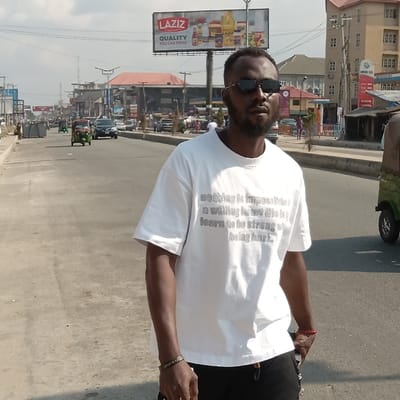 A man wearing sunglasses and a white t-shirt with printed text stands outdoors. The t-shirt reads "Nothing is impossible... a willing to not the learn to be strong so being hard." A "LAZIZ QUALITY YOU CAN TRUST" billboard is visible in the background, along with a street scene that includes buildings, vehicles, and pedestrians. The image is taken in daylight with a slight tilt.