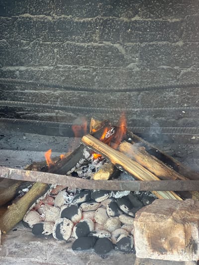 The image shows a close-up view of a fire burning in a barbecue pit. The fire is composed of logs and charcoal briquettes, with flames visible. The pit itself appears to be constructed of rough, textured brickwork, possibly in an outdoor setting. There is a metal grate or support running across the middle of the pit, separating the burning material from the lower section where more charcoal is visible. The background consists of the smoky, textured interior walls of the barbecue pit. No people are present in the image.