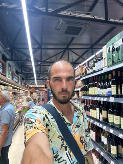 A man is taking a selfie in a supermarket aisle. He is in the foreground, and his face is partially visible. He has short dark hair and a beard, and he is wearing a colorful, patterned shirt. A dark blue strap from a bag is visible across his chest. Behind him, shelves are stocked with various items, including bottles of wine. Other shoppers are visible in the background, pushing shopping carts. The supermarket appears well-lit with overhead fluorescent lighting. The scene is likely within Ulcinj - Ulqin, Montenegro, as indicated by the user.