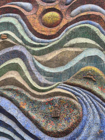 This image shows a close-up view of a colorful mosaic wall art located in the city of Borjomi, Georgia. The mosaic depicts a stylized landscape with wavy bands of various colors, suggesting hills or water. Dominating the upper portion of the mosaic is a large, circular sun or celestial body rendered in shades of orange, red, and yellow.
Scattered throughout the mosaic are small, three-dimensional bronze sculptures. In the upper left, there is a miniature house. In the middle right, a small bronze deer is depicted mid-leap across a textured area. In the lower left, a miniature church or chapel stands on a section of mosaic filled with small, colorful floral patterns. In the lower right, a small bronze fish is visible. The mosaic itself is composed of numerous small, irregularly shaped tiles in a wide range of colors, including blues, greens, browns, reds, oranges, and yellows, creating a vibrant and textured surface. The overall impression is of a whimsical and artistic representation of nature and small settlements.