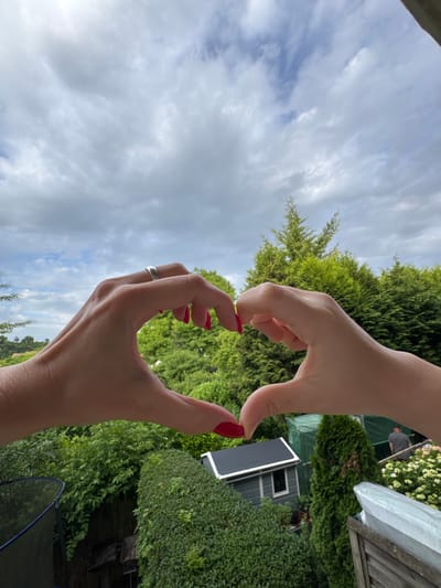 The image shows two hands forming a heart shape against a cloudy sky and a lush green backyard. The left hand has a silver ring on the index finger and bright red nail polish. The right hand also has bright red nail polish. In the background, there are numerous evergreen trees and hedges, a small gray shed with a black roof, a greenhouse, and a wooden fence. A person wearing a dark gray t-shirt is visible in the distance, standing near the greenhouse. The overall scene suggests an outdoor setting, likely a residential backyard. The location is Bietigheim-Bissingen, Germany.