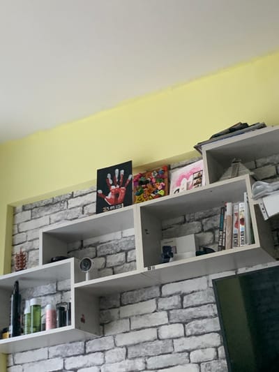 The image depicts a cluttered white shelving unit mounted on a gray brick-patterned wall. The shelves are adorned with various objects, including bottles of toiletries, books, a small camera, and several framed artworks. One artwork is a child's handprint painting dated 05.05.2019. Another appears to be a collage of colorful pom-poms. On the top shelf, a stack of magazines or papers is visible, along with a miniature pyramid model. The shelving unit is positioned below a yellow and white ceiling. The overall scene suggests a domestic setting, possibly a living room or bedroom, in Severodvinsk, Russia.