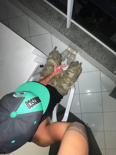 A person sits on a white chair, wearing camo pants and gray sneakers. Their left hand displays a three-finger gesture with a small, circular object between the fingers. The person is wearing a black t-shirt and a black baseball cap with green details. The person's legs are tattooed. They are sitting on a tiled floor near a stone structure.