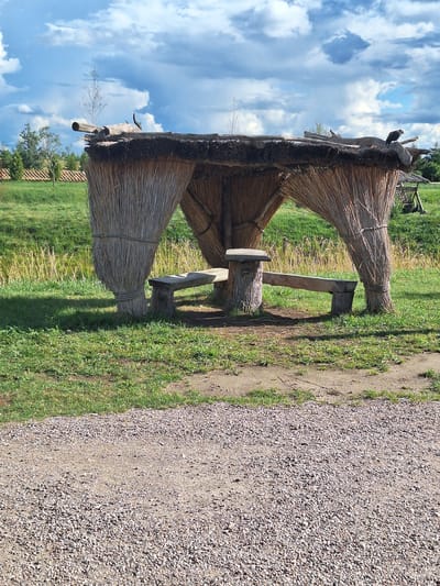 The image depicts an outdoor scene with a thatched gazebo constructed from straw and wood, featuring two rustic wooden benches and a central log table. The gazebo is situated in a grassy area adjacent to a path made of gravel and dirt. In the background, there is a body of water, lush green vegetation, and trees. The sky is partly cloudy with a mix of blue sky and white clouds. The overall setting suggests a natural, rural environment.