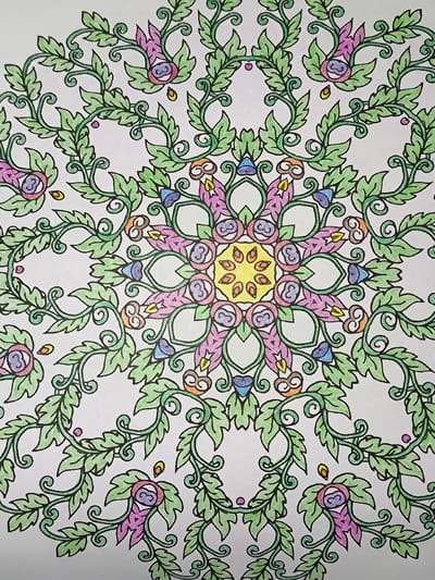 The image is a close-up, top-down view of a section of a mandala coloring page. The mandala features a symmetrical floral and vine design. The dominant color is green, used for the swirling vines and numerous leaves. Interspersed throughout the design are small, petal-like shapes in shades of purple and pink, some with blue accents. At the center of the visible portion of the mandala is a yellow, star-like shape. There are also small details in orange and purple within the design. The coloring is somewhat uneven, indicating it is a hand-colored page. The background is white, suggesting the image is printed on paper. There is no indication of any real-world location, including the city of Pomezia, Italy. The image depicts a static, artistic creation.