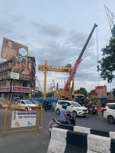 This image captures a bustling street scene in Chennai, India, dominated by a large construction project. In the foreground, yellow barricades line the road, displaying traffic rules and advertisements for Apollo Hospitals. Several vehicles, including cars and motorcycles, are seen on the asphalt, indicating moderate traffic. A man in a blue shirt and helmet is riding a scooter, suggesting daily commute activities.
The background is dominated by a massive construction crane, its red and yellow boom extending high into the cloudy sky. Another crane, yellow and brown, is also visible, hinting at ongoing infrastructure development. The sky is overcast, suggesting a cloudy day, possibly during the monsoon season or a period of heavy cloud cover.
Advertisement billboards add color and information to the scene. One billboard prominently features "GRT JEWELLERS" and a woman in traditional attire. Another advertises "BHIMA" jewelry and "Hot Breads." The text on the billboards is a mix of English and Tamil, common in Chennai. The presence of construction machinery, traffic, and advertisements collectively paints a picture of a dynamic urban environment in Chennai. The overall impression is one of a city in motion, with development and daily life coexisting.