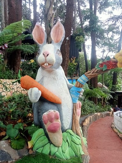 The image displays a large, prominently featured rabbit statue, situated outdoors in Kuwukan, Indonesia. The statue is grey and white with pink interior ears, nose, and paw pads, depicted in a sitting pose. It holds a large orange carrot with a green top in its left hand. Its right arm is wrapped with blue and white striped bandages, gripping a brown, gnarled staff adorned with red flowers and yellow foliage/fruit at its top. The statue rests on a sculpted base resembling green leaves, surrounded by artificial green turf and natural broadleaf plants and ferns. Behind the statue on the left, a low, light-colored wall with a textured pattern is visible. A red-brown paved pathway curves from the midground towards the distant right background. This path is bordered by a low white picket fence on its right and a low, wood-textured curb on its left. The background is dominated by numerous tall coniferous trees and dense green undergrowth. A dark building structure is partially visible among the trees on the left. A solitary human figure, dressed in dark clothing, is faintly discernible on the pathway in the far background.