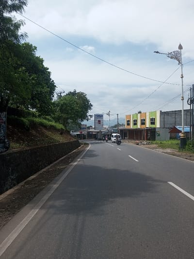 The image displays a daytime outdoor scene on a paved two-lane road in the city of Chhenangri_Rishor, Bhutan, under a partly cloudy sky. On the left, an elevated, tree-covered embankment is retained by a dark grey stone wall. On the right side of the road, a series of structures includes a partially constructed building with exposed concrete walls, followed by a commercial building featuring a facade with alternating orange, green, and light yellow vertical panels, fitted with multiple square windows and an open ground floor. Further down the road, a large white billboard with blue text is visible. A decorative white street lamp post with gold accents and an ornate finial stands on the far right. Utility poles and overhead lines are present throughout the scene. Three vehicles are on the road, traveling away from the viewer towards the background: a white minivan and two motorcycles. Distant hills or mountains are visible in the background. No specific individuals are discernible on foot.