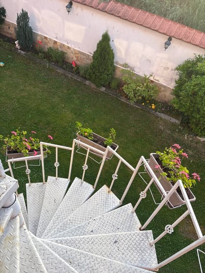This is an overhead, high-angle shot of a spiral staircase made of metal with a diamond plate pattern. The staircase is painted white and has a decorative railing with a swirling motif. On the landing at the top of the stairs are two planters filled with pink flowers. The staircase overlooks a lush green lawn. To the left of the lawn, there is a paved walkway with a black lamp post and a section of red brick wall or edging. There is also a small evergreen tree and a garden bed with some red flowers. The scene appears to be outdoors, likely in a garden or yard, during daylight. The lighting suggests it might be morning or late afternoon due to the soft shadows. The location is specified as Strelcha, Bulgaria. There are no people visible in the image.