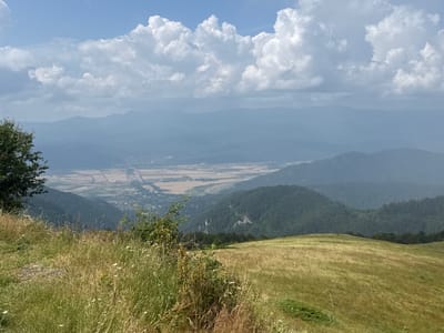 The image shows a wide, panoramic view of a mountainous landscape under a cloudy sky. In the foreground, a grassy hillside slopes downwards, with dry, golden grasses and some scattered green bushes and small white flowers. To the left, a dark green tree stands tall.
The midground features rolling, forested hills covered in dark green trees. Between these hills, a valley opens up, revealing agricultural fields with a patchwork of light brown and tan colors, suggesting crops ready for harvest. A few buildings are visible in the distance within the valley.
The background consists of several layers of misty, blue-hazed mountains, diminishing in clarity with distance. The sky is filled with large, white cumulus clouds, some appearing bright and fluffy, others darker and more diffused, set against a pale blue sky.
The overall impression is of a vast, natural landscape, likely a rural area. Based on the provided context, this scene is located in or near the city of Hristo Danovo, Bulgaria.